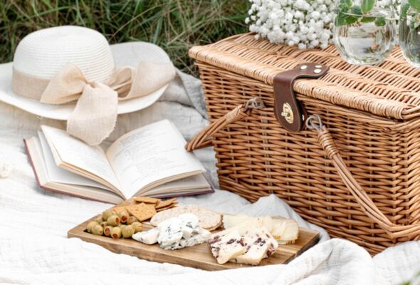 Ein Picknickkorb vom Restaurant Gut Ising am Chiemsee steht auf einer Wiese, hergerichtet mit Picknickdecke, Sonnenhut und einem Buch zum Lesen.